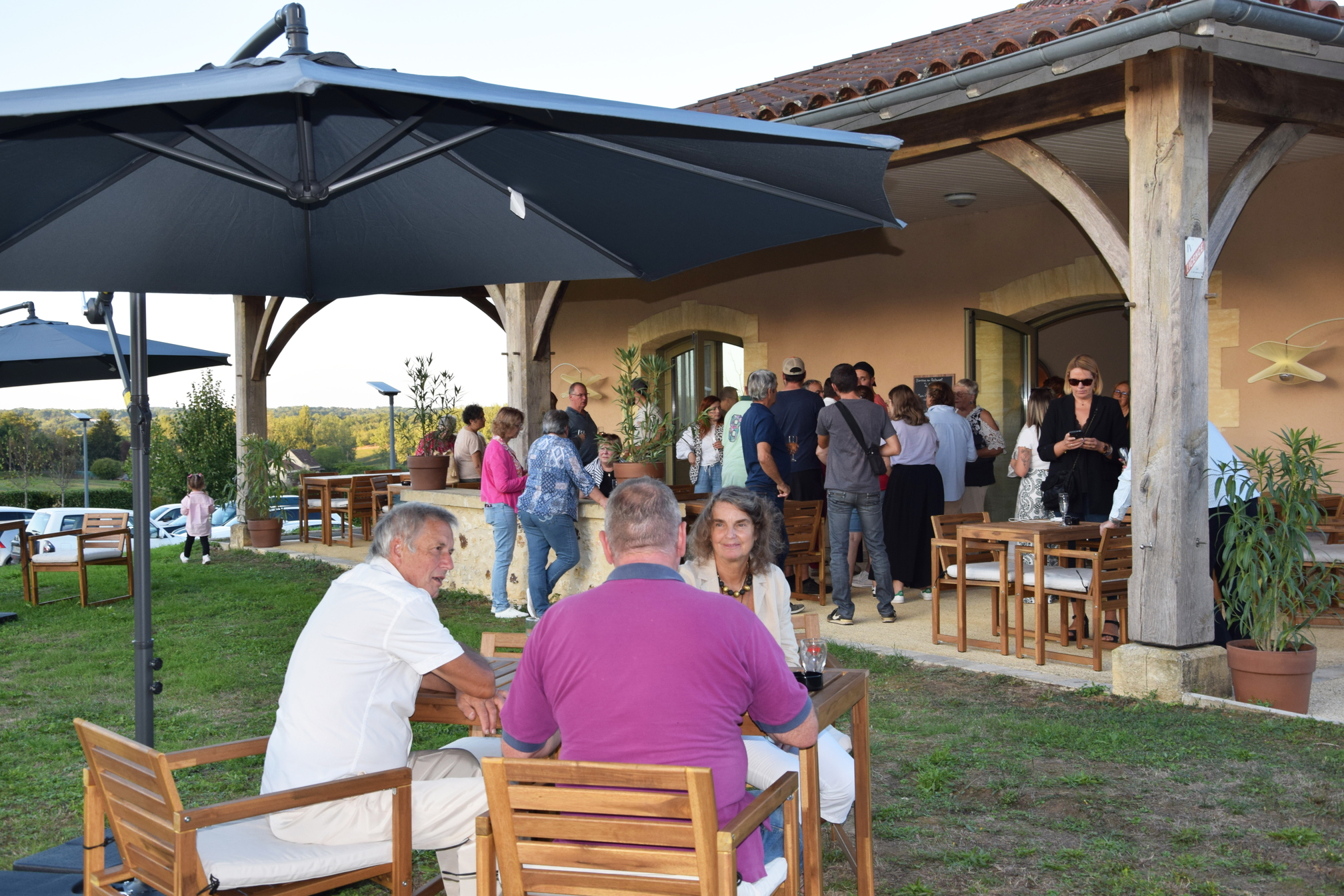 Des personnes sont assises à une table, sous un parasol. Elles font face à une terrasse où les personnes sont debout, un verre à la main. Elles participent à l'inauguration du nouveau restaurant Le Cendrieux.