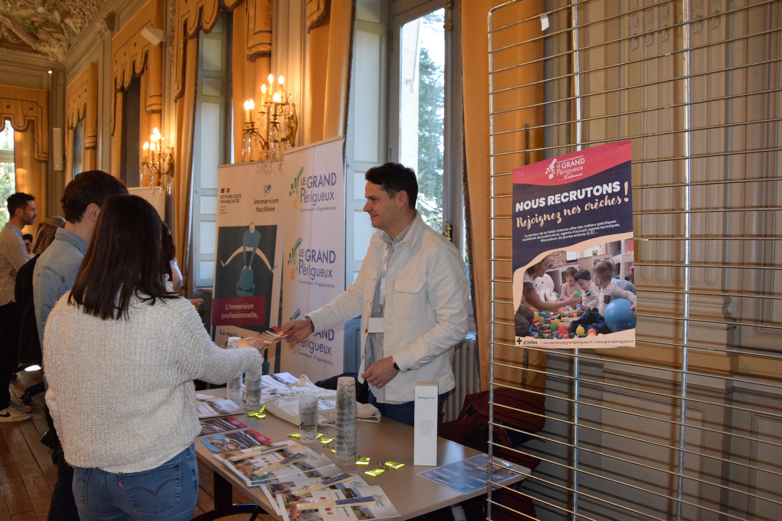 Un homme tenant un stand de recrutement et donnant un prospectus à deux personnes venues se renseigner sur les offres d'emploi. On aperçoit une affiche avec écrit "Nous recrutons, rejoignez nos crèches !"