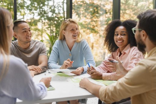 Des jeunes souriants sont assis à table. Ils échangent et communiquent entre eux. Une femme blonde porte un pull bleu, une femme aux cheveux bruns, crépus, porte une veste rose.