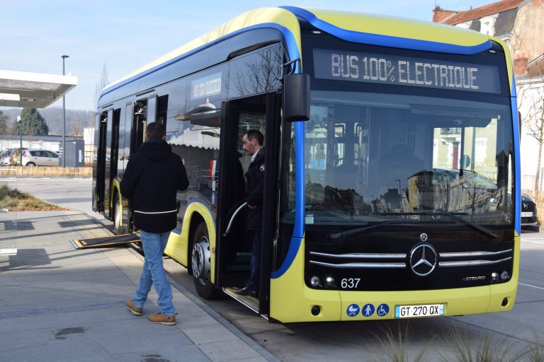 un bus jaune avec plusieurs bandes en bleu est stationné. A l’avant du bus, un homme descend par la porte et une autre personne marche à côté du bus. Sur le devant du bus, il est écrit en gros : bus 100% électrique