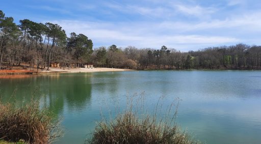 L'étang de Neufont, avec sa couleur oscillant entre le bleu et le vert et des arbres autour. Au fond, sur une petite plage, la guinguette