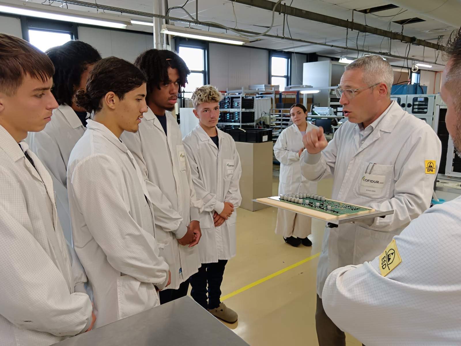Groupe de jeunes dans l'entreprise industrielle Cofidur. Ils portent des blouses blanches. Un homme plus âgé, lui aussi portant une blouse blanche, montre une carte électronique, le savoir-faire de l'entreprise.