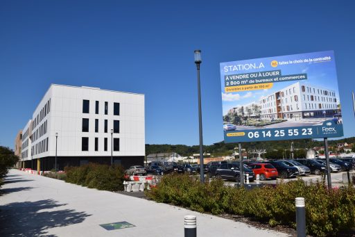 Sous un ciel bleu très vif, au 1er plan, à l'entrée du quartier Aliénor, marquée par des bâtiments, le long d'un chemin, et un parking avec des voitures, un grand panneau indique Station A, à vendre ou à louer. 2800m2 de bureaux et commerces