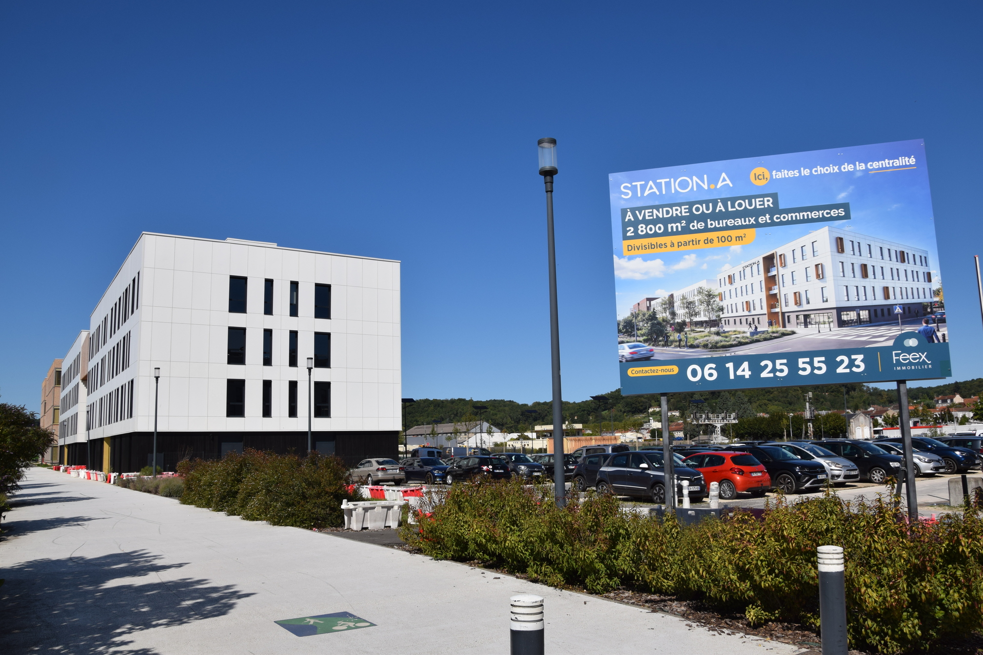 Sous un ciel bleu très vif, au 1er plan, à l'entrée du quartier Aliénor, marquée par des bâtiments, le long d'un chemin, et un parking avec des voitures, un grand panneau indique Station A, à vendre ou à louer. 2800m2 de bureaux et commerces