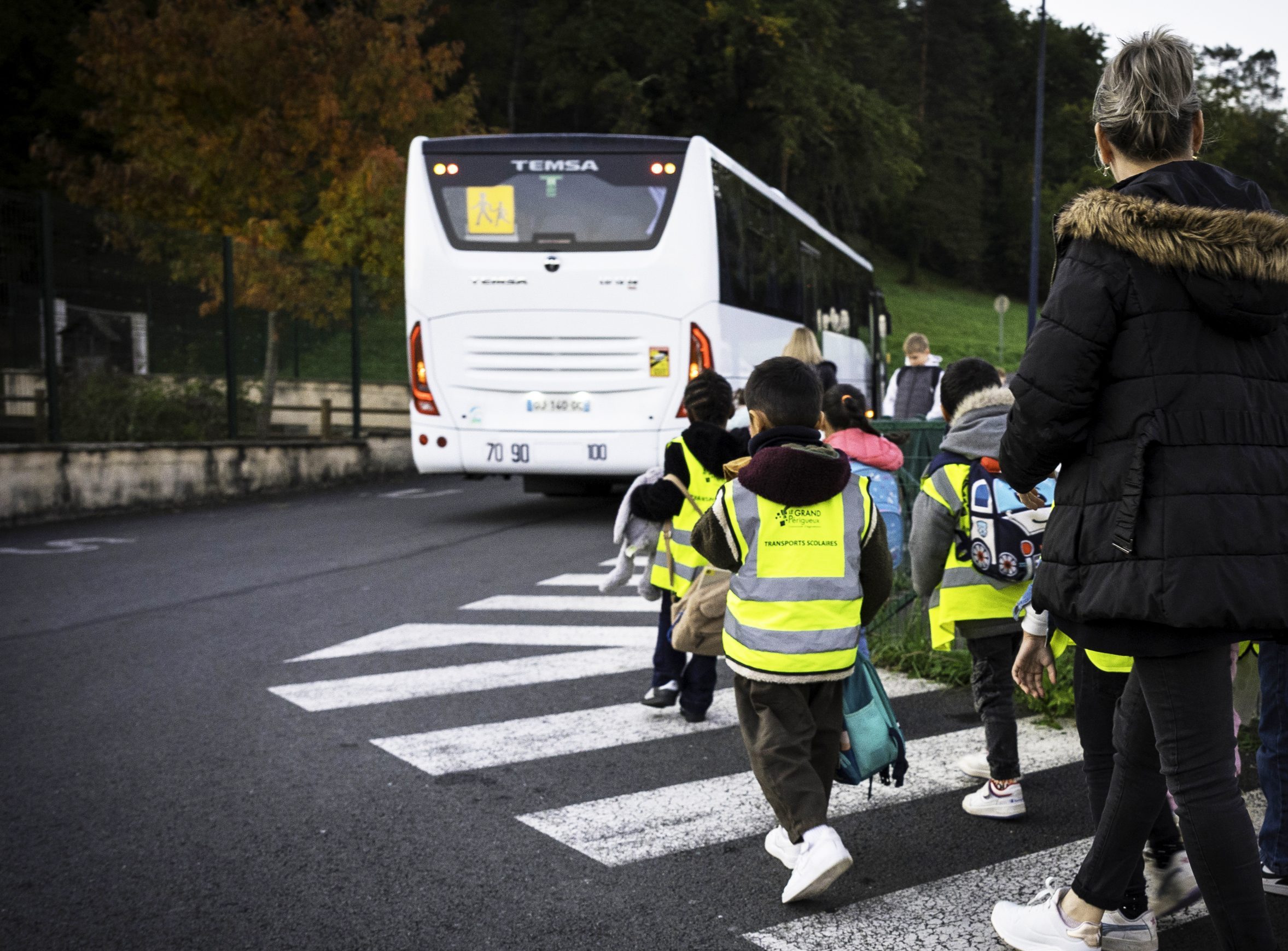 Des enfants de dos portent des brassards fluos inscrits Grand Périgueux. Ils vont prendre un autocar