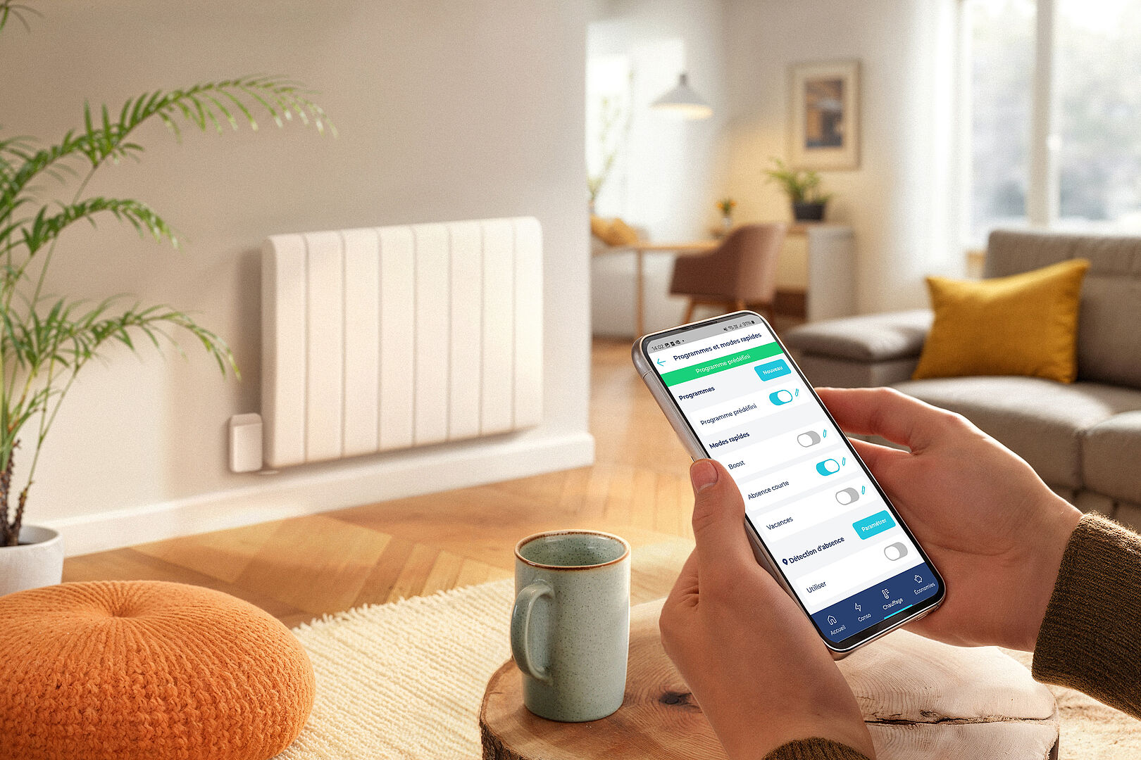 Dans un salon cocooning, aux couleurs beige et marron, une main tient un téléphone portable, à côté d'une tasse et surtout en face d'un radiateur électrique