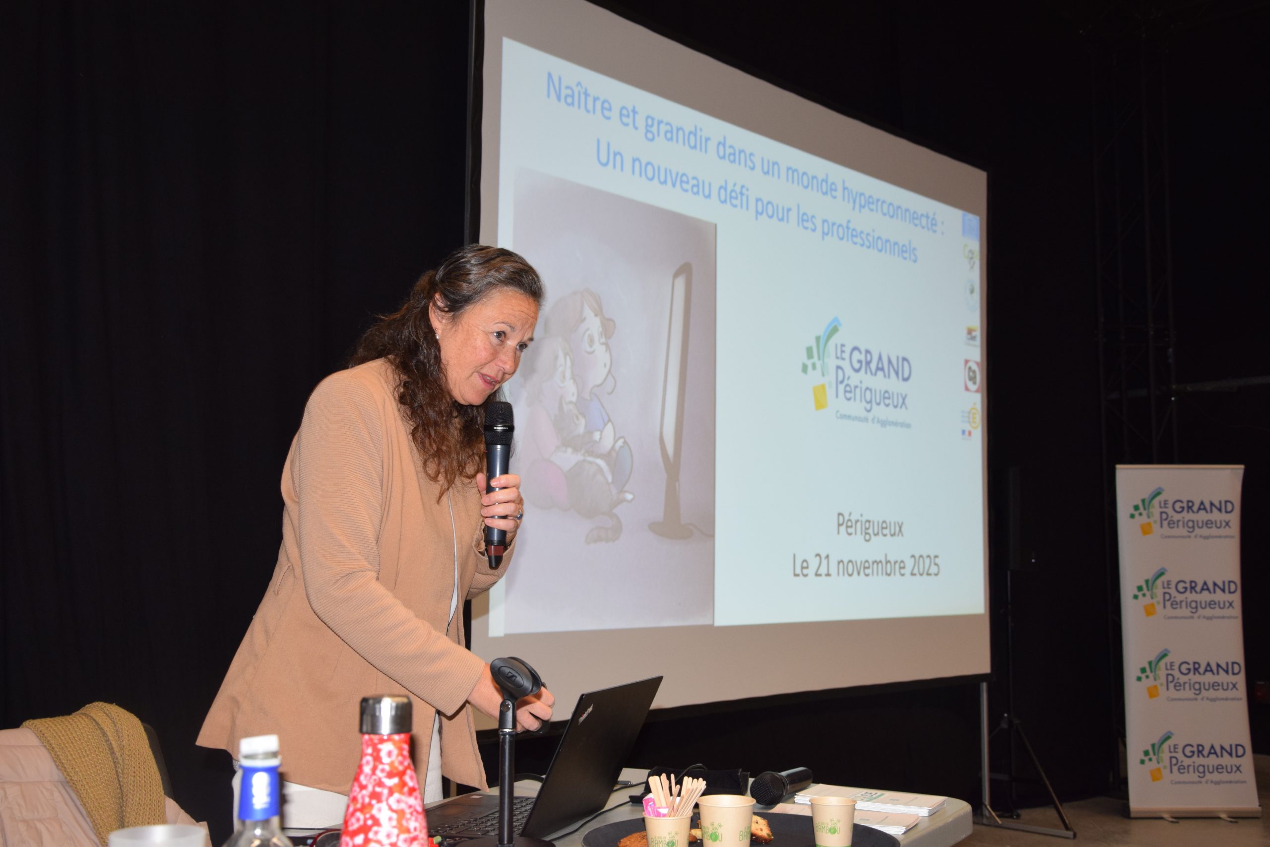 Une dame avec une veste beige, s'adresse à un public. Derrière elle, sur l'écran, il est écrit "Naître et grandir dans un monde hyperconnecté : un nouveau défi pour les professionnels.