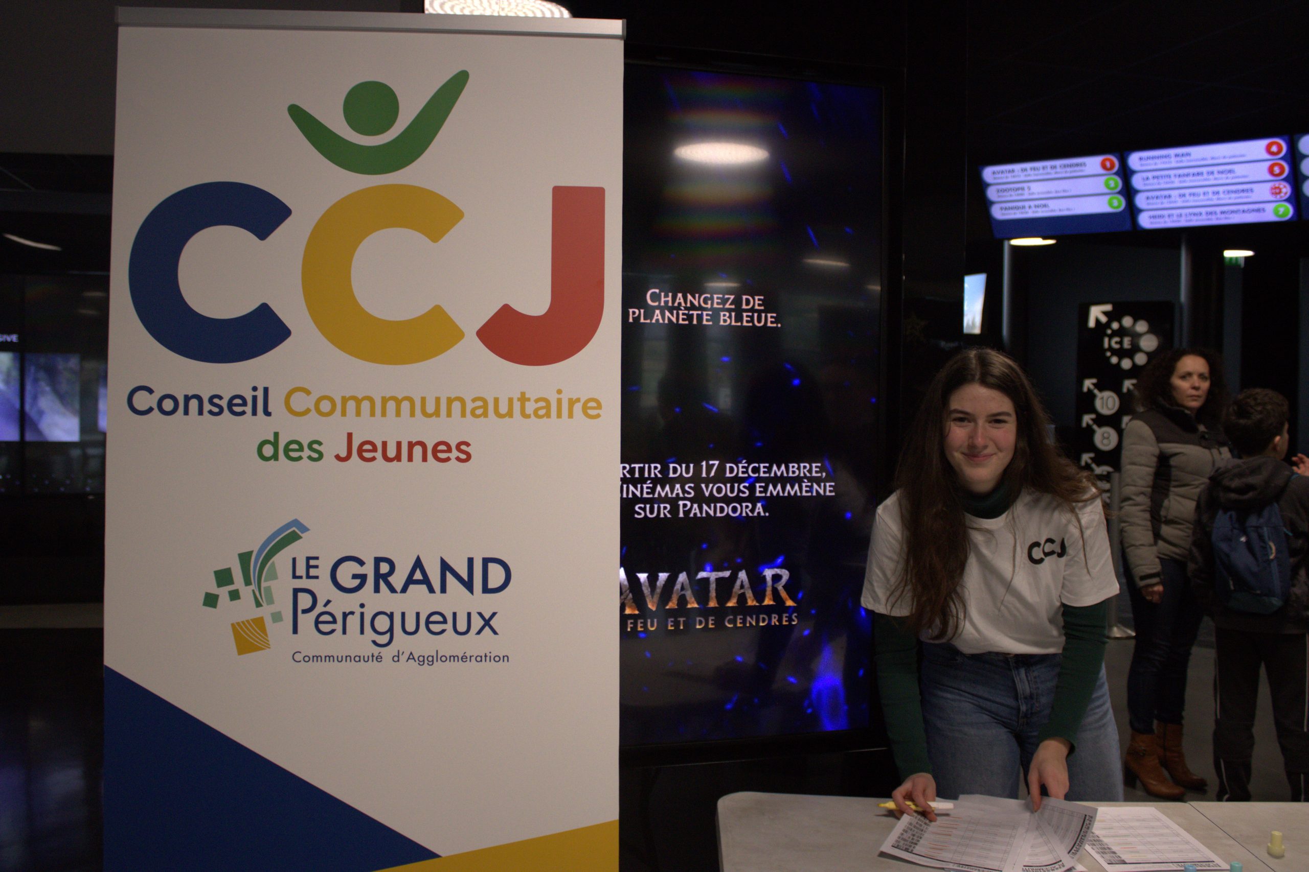 Une des membres du conseil communautaire des jeunes du Grand Périgueux tient un stand pour en faire la promotion dans le hall du cinéma CGR de Périgueux. En arrière plan on peut voir l'affiche du film Avatar que les jeunes sont allés voir.
