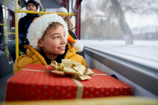 Un enfant avec un anorak jaune et un bonnet style bonnet de Noël tient un paquet sur lui. Il est dans un bus.