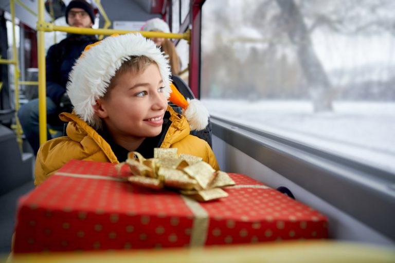Un enfant avec un anorak jaune et un bonnet style bonnet de Noël tient un paquet sur lui. Il est dans un bus.