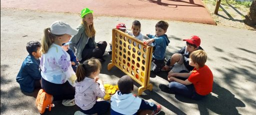 Une animatrice avec une casquette verte encadre des enfants jouant avec un panneau en bois comprenant des espaces pour passer des boules