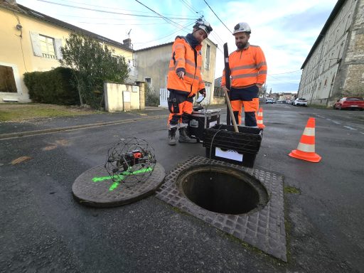 Deux hommes en combinaisons de travail de couleur orange fluorescent et munis de casques se trouvent dans une rue à proximité d'un regard d'eaux fluviales. À leurs pieds se trouvent un drone équipé d'une cage de forme ronde ce qui permet à l'engin de volet dans des réseaux sous terrain et de ne pas être endommagé s'il percute une paroi.