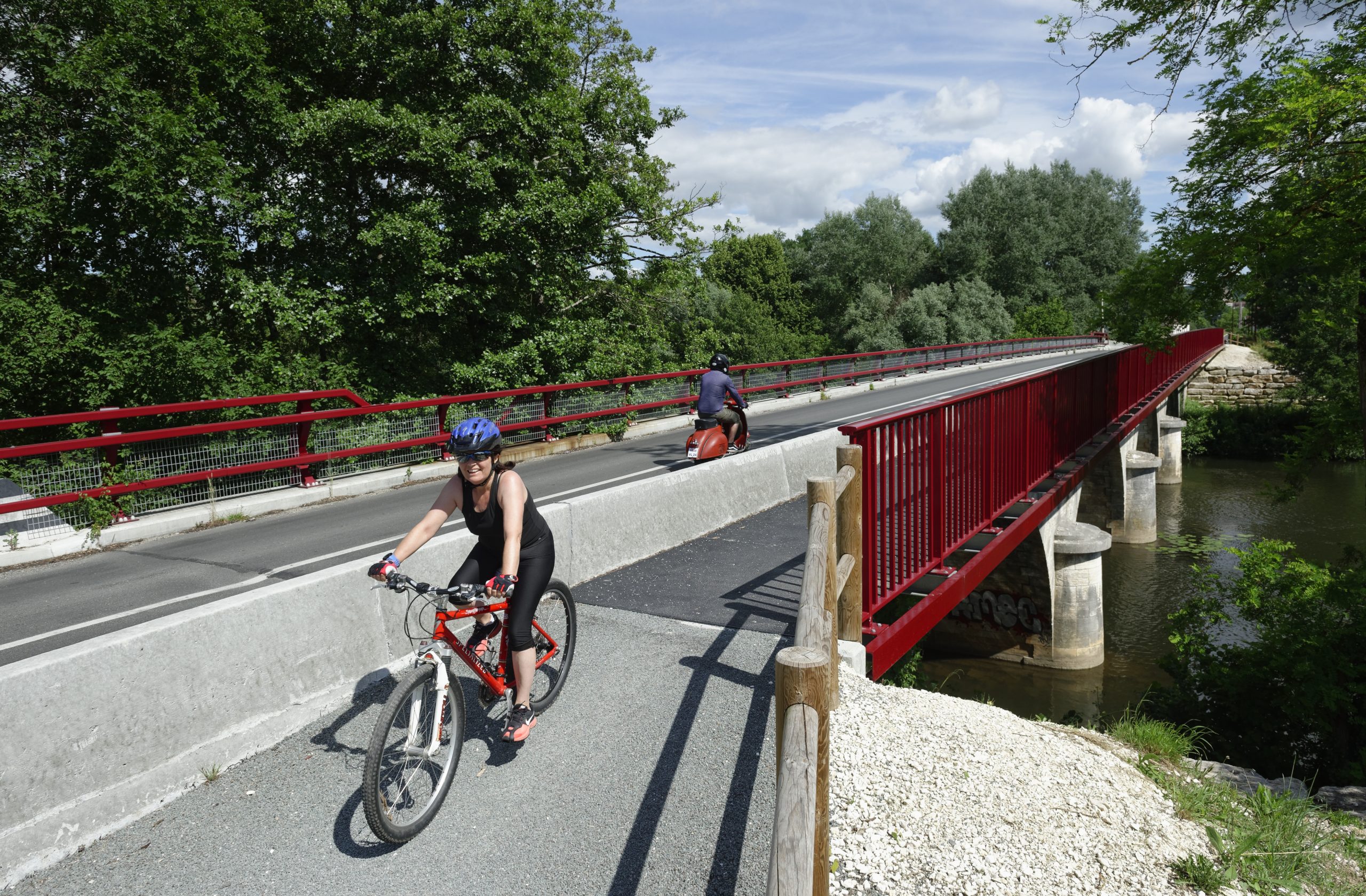 Un cycliste habillé en tenue estivale et en noir circule sur la sortie d'une route près d'une passerelle dont les bords sont peints en rouge. Près des piliers, coule la rivière.