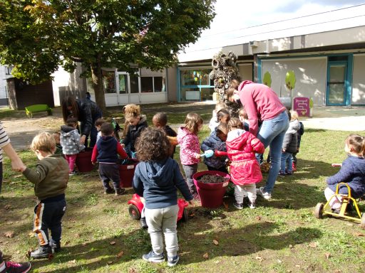 Un groupe d'enfants habillés chaudement jouent à l'extérieur. Ils sont entourés d'un adulte, en jean bleu et pull rose.
