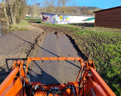 La voie verte est en cours de nettoyage après les inondations. Une longue portion de boue marron avec des sédiments a remplacé la voie. Un engin de couleur orange procède au nettoyage pour enlever la boue
