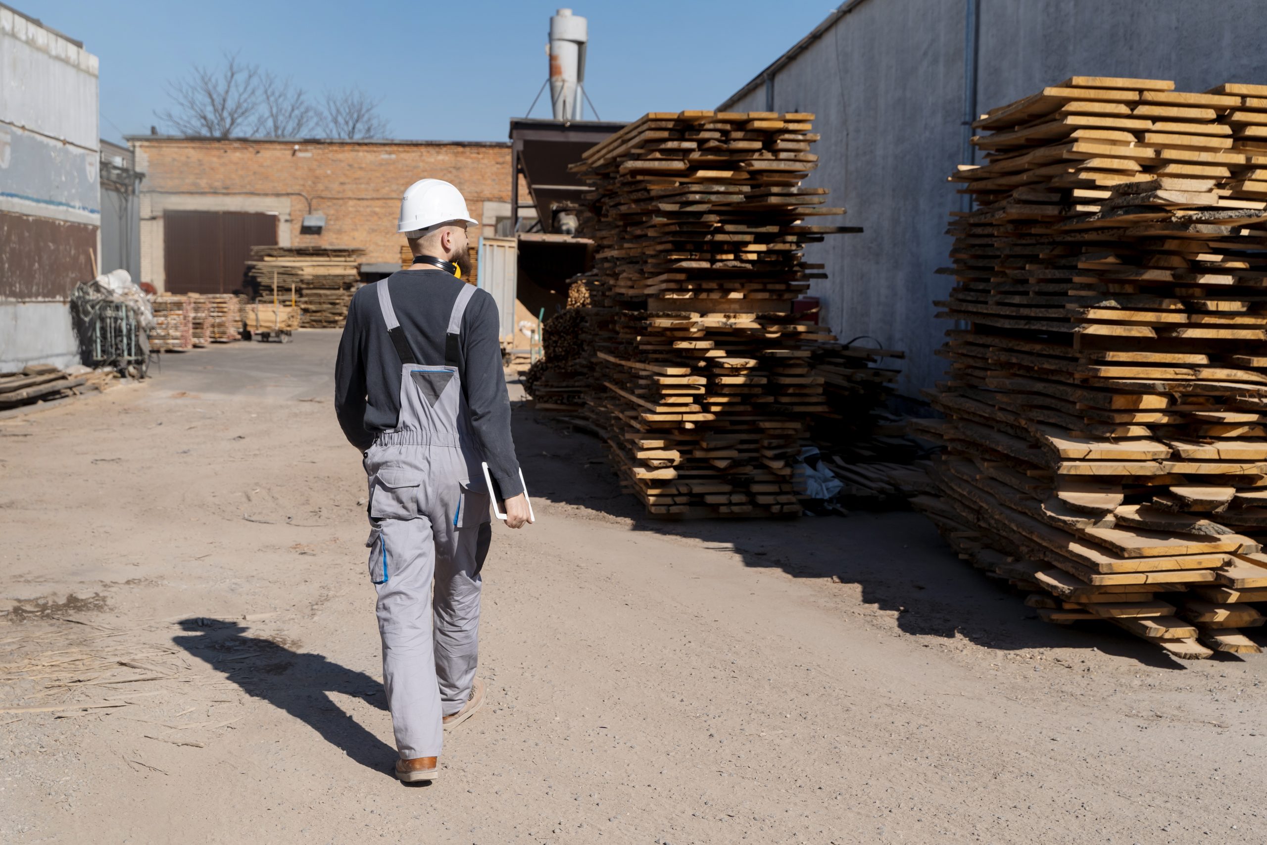 Un homme vêtu d'une salopette grise du bâtiment et portant un casque du BTP regarde à droite un gros tas de bois et de palettes.