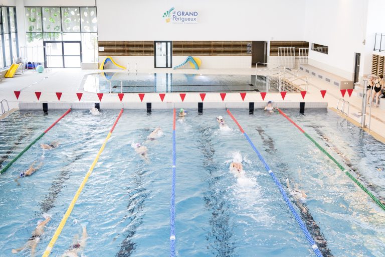 Sur plusieurs couloirs de nage dans un grand bassin, des personnes pratiquent la natation. Au fond, le petit bassin et des petits toboggans.